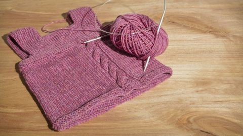 Ein pinkfarbener Strickpullover liegt flach auf einem Holzuntergrund neben einem Knäuel Wolle und Rundstricknadeln.