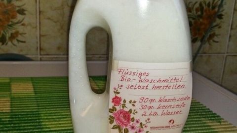 Eine Flasche mit flüssigem Waschmittel steht auf einer Waschmaschine. Die Küche ist mit Blumenmustern dekoriert.