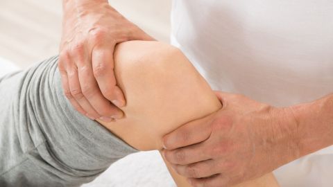 Ein Therapeut massiert sanft ein schmerzhaftes Knie, um Knieschmerzen zu lindern, in einem hellen, neutralen Raum.