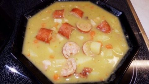 Eine sämige Erbsensuppe liegt in einer schwarzen Schüssel, umgeben von Wurststücken und Gemüse, auf einem Herd.