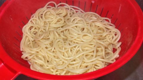 Spaghetti liegen in einem roten Sieb und warten darauf, abgetropft zu werden, während der Küchenbereich im Hintergrund sichtbar ist.