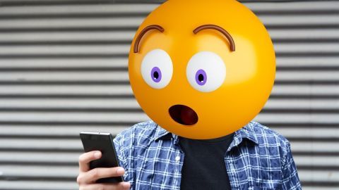 Ein erstaunter Emoji-Kopf sieht auf ein Smartphone, während er in einem urbanen Umfeld steht, umgeben von einer gestreiften Wand.