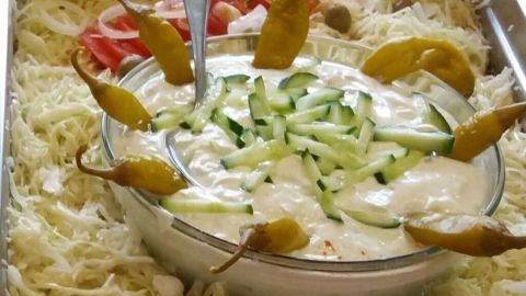 Eine Schüssel Zaziki ist mit Gurkenstücken und Gewürzgurken garniert, umgeben von frischem Gemüse wie Salat, Tomaten und Zwiebeln.