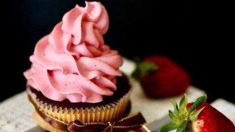Ein Muffin mit Erdbeer-Vanillepudding-Frosting ist liebevoll dekoriert und steht neben frischen Erdbeeren auf einer Holzoberfläche.