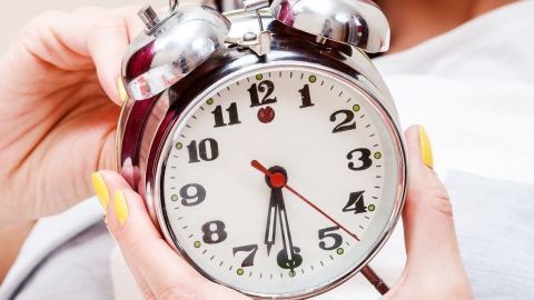 Eine Frau hält einen Wecker in der Hand, während sie im Bett liegt, um ihren Körperrhythmus auf Sommerzeit einzustellen.