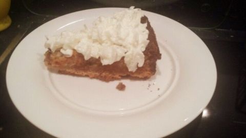 Ein Stück cremiger Bananen-Schokoladenkuchen liegt auf einem weißen Teller, garniert mit Sahne. Der Kuchen steht in einer Küche.