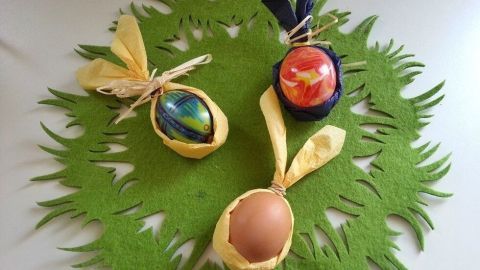 Bunte Ostereier, eingewickelt in gelbe Servietten, liegen auf einem grünen Filzuntergrund und dekorieren fröhlich den Tisch.