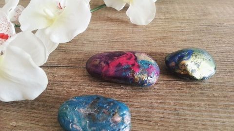 Bunte Dekosteine mit Nagellack liegen auf einem Holztisch, umgeben von weißen Orchideenblüten und frischem Grün.