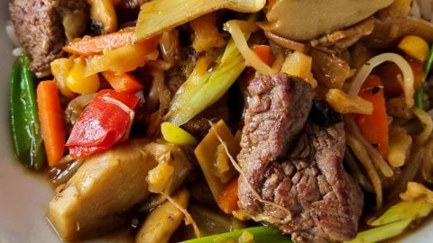 Zartes Filetstück liegt in aromatischer Sojasoße, umgeben von buntem Gemüse und Nudeln auf einem weißen Teller.