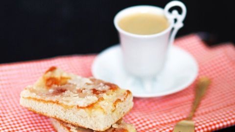 Zwei Stück Zuckerkuchen liegen auf einem karierten Tuch neben einer Tasse Kaffee auf einem schwarzen Tisch.