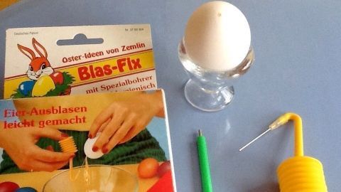 Ein Ei steht auf einem Eierbecher, umgeben von einem Blasgerät und einer Anleitung für das einfache Eier Ausblasen.