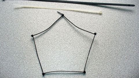 Ein Kabelbinder ist in einem fünfeckigen Rahmen angeordnet, während ein gerader Stab daneben liegt, auf einer hellen Oberfläche.