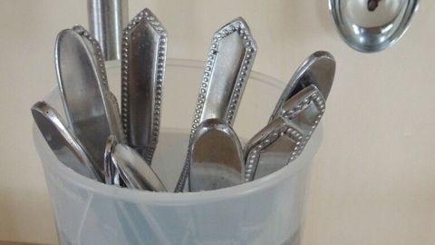 Es passiert immer wieder, dass Besteck im Geschirrspüler Flecken bekommt oder Rost ansetzt. Wie kann man das vermeiden? Edelstahlbesteck steht in einem Glas mit Wasser, um Flecken zu vermeiden, auf einer Holzoberfläche in einer Küche.
