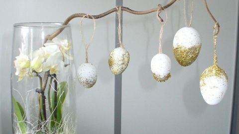 Ein eleganter Ast mit hängenden Ostereiern in einer Vase, dekoriert mit weißen Blumen und Gräsern, schafft einen glamourösen Ostergruß.