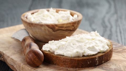 Eine Holzschale mit frisch zubereitetem Quark steht neben einer Scheibe Brot, die ebenfalls dick mit Quark bestrichen ist.