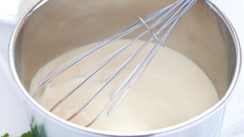 Eine silberne Schüssel enthält eine cremige Sauce Hollandaise, während ein Schneebesen darin rührt. Im Hintergrund sind Butter, Ei, Zitrone und frische Kräuter zu sehen.
