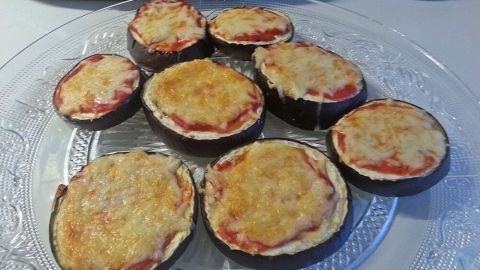 Kleine Auberginen-Pizzen liegen auf einem Glasservierplatte, überbacken mit Käse und Tomatensauce, daneben liegt eine Zucchini.