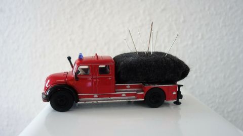 Ein rotes Spielzeugauto mit einem schwarzen Nadelkissen auf der Ladefläche steht auf einem weißen Tisch vor einer Wand.