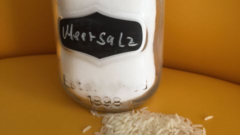 Ein Glas mit Meersalz steht auf einer gelben Fläche, während etwas Reis daneben verstreut ist.