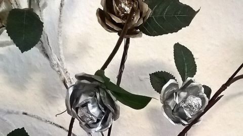 Silberne Rosen aus Teelichtschalen stehen in einer weißen Vase, umgeben von grünen Blättern und Zweigen.