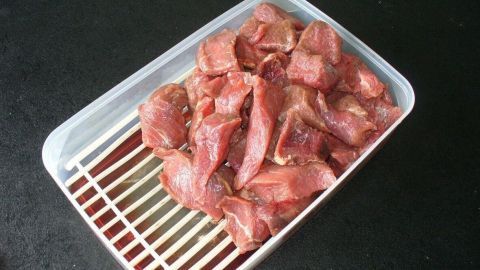 Rohes, gefrorenes Fleisch liegt in einer transparenten Schale mit einem Gitterboden auf einer dunklen Oberfläche.