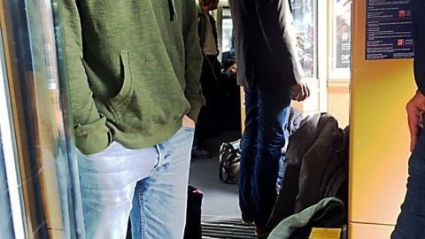 Eine Gruppe von Fahrgästen steht dicht gedrängt im überfüllten Zug, während andere im Hintergrund sitzen und die Einfahrt beobachten.