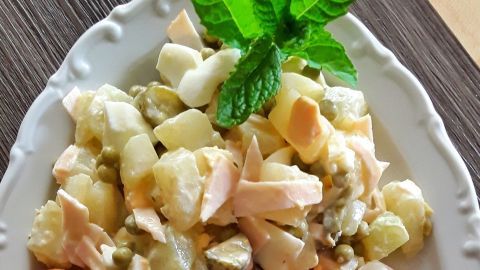 Ein bunter Kartoffelsalat liegt auf einem weißen Teller neben zwei Würstchen und Tomatenscheiben, garniert mit Minze.