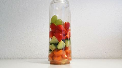 Ein transparenter Smoothie Mixer ist gefüllt mit bunten Früchten wie Melonen, Trauben und Gurken, vor einem neutralen Hintergrund.