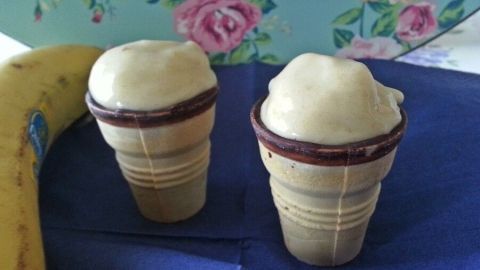 Zwei Eiswaffeln mit Bananeneis stehen auf einem blauen Untergrund, neben einer Banane, vor einem floral gemusterten Hintergrund.