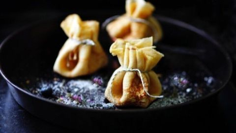 Pfannkuchen mit der Zitronencreme füllen, in kleine Säckchen formen und mit den Vanilleschotenhälften oder Paketband zubinden. Drei gefüllte Pfannkuchen-Zitronen-Säckchen liegen auf einer schwarzen Pfanne, umgeben von Puderzucker.