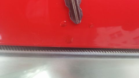 Eine rote Autokarosserie zeigt einen kleinen Steinschlag in der Lackierung mit metallischem Hintergrund.