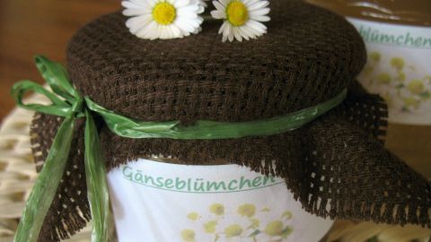 Ein Glas Gänseblümchen-Gelee ist hübsch dekoriert mit einem braunen, gehäkelten Deckel und verziert mit zwei echten Gänseblümchen.