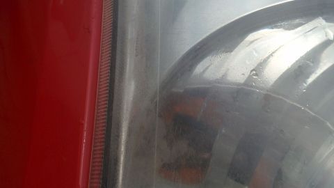 Trübe Scheinwerfer einer roten Fahrzeugfront reflektieren schwaches Licht, umgeben von einer unscharfen Umgebung.