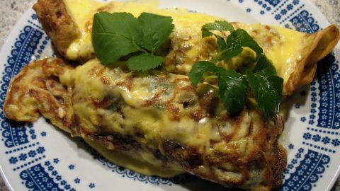Zwei überbackene Schinken-Spargel-Crepes liegen auf einem blauen Teller mit einem schönen Muster und sind mit Kräutern garniert.