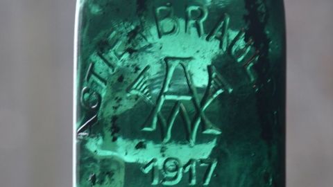 Eine grüne Bierflasche mit Rostflecken steht im Fokus, beleuchtet von sanftem Licht in einer neutralen Umgebung.