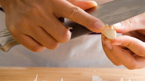 Hände schälen eine Knoblauchzehe, während zwei ganze Knoblauchköpfe und Schalen auf einem Holzbrett liegen.
