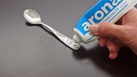 Ein Löffel wird mit Zahnpasta gereinigt. Die Hand hält die Zahnpastatube über den Löffel, auf einem dunklen Tisch.