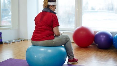 Eine Person sitzt auf einem blauen Gymnastikball in einem hellen Raum mit Fitnessgeräten und bunten Bällen im Hintergrund.