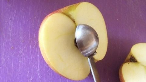 Ein halbierter Apfel liegt auf einer lila Schneidefläche, während ein Löffel das Kerngehäuse schnell entfernt.