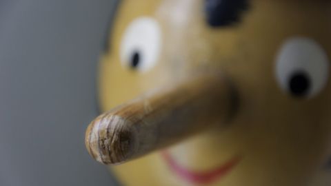 Ein Holzspielzeug mit einer langen Nase lächelt neugierig in einem grauen Hintergrund, symbolisiert die Idee, dass Lügen kurze Beine haben.