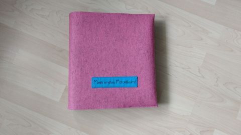 Eine pinke Filzhülle schützt ein Fotoalbum. Sie liegt auf einem hellen Holztisch und hat ein blaues Etikett.