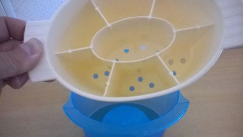 Ein gelber Besteckkorb wird handgehalten über einem blauen Behälter, bereit zur Reinigung in der Spülmaschine.
