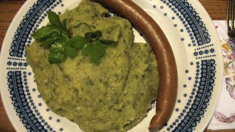 Grünes Kartoffel-Kohlrabi-Püree, das auf einem Teller serviert wird, zusammen mit einer Würstchen und frischen Kräutern.