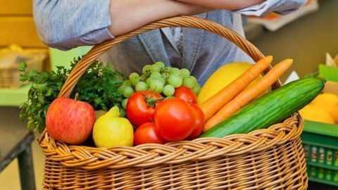 Eine lächelnde Frau lehnt sich an einen Korb voller frischem Obst und Gemüse in einem hellen, einladenden Markt.