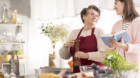 Zwei Frauen in einer hellen Küche bereiten gemeinsam ein Gericht vor, während eine das Rezept in einem Buch liest.