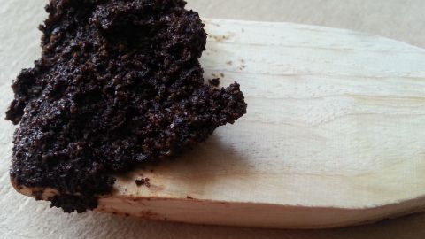 Ein Holzlöffel hält frischen Kaffeesatz, der als Cellulite-Peeling genutzt wird, auf einem neutralen Hintergrund.