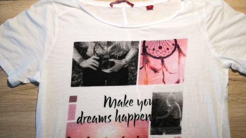 Eine weiße T-Shirt mit einem bunten Druck, das auf einem Holztisch liegt, zeigt den Text „Make your dreams happen“.