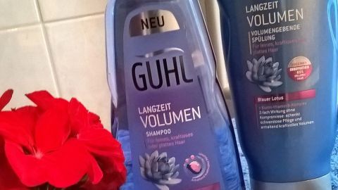 Shampoo und Spülung von Guhl für längeres Volumen stehen neben einer roten Blume auf einem blauen Handtuch im Badezimmer.