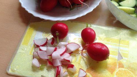 Frische Radieschen liegen auf einem Tisch, während einige bereits in Scheiben geschnitten sind. Neben ihnen ist eine Schüssel mit Gurken.