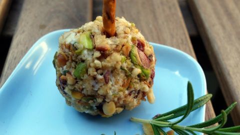 Eine Käse-Pop-Kugel auf einem blauen Teller, umgeben von gehackten Nüssen und frischen Kräutern, auf einem Holztisch.